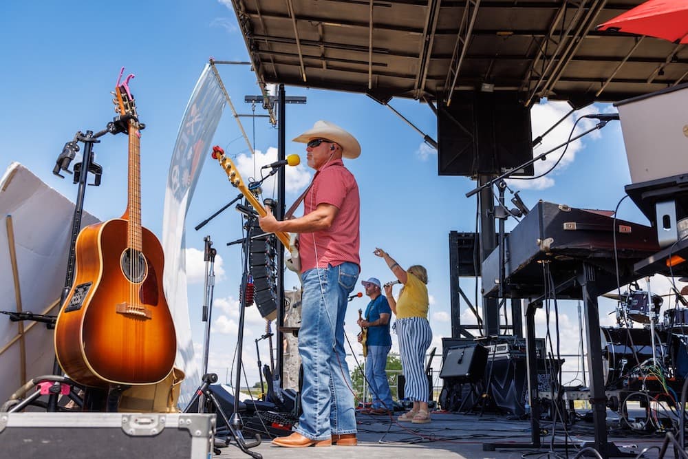 A band performs live on an outdoor stage at Mayfest 2025; the guitarist sings into a microphone while other musicians and instruments are visible in the background.