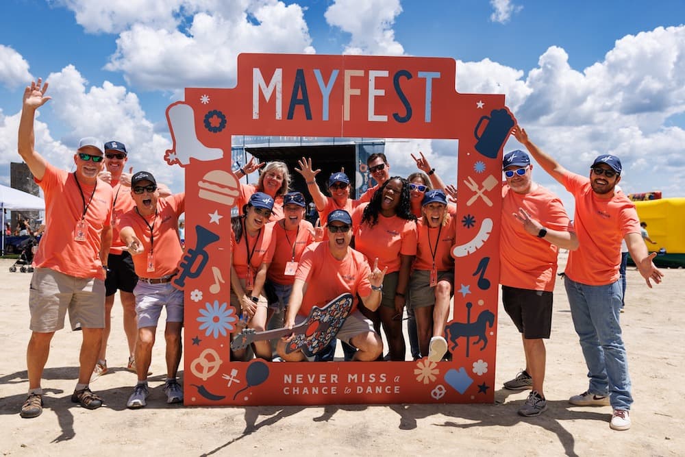 Southstar staff dressed in matching orange shirts pose and smile behind a large "Mayfest" photo frame at an outdoor event under a blue sky with clouds.