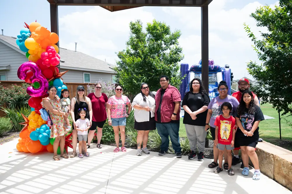 A group of people pose for a photo outdoors under a pergola at the VIDA San Antonio development, with balloons shaped as the number three and a bounce house visible in the background.