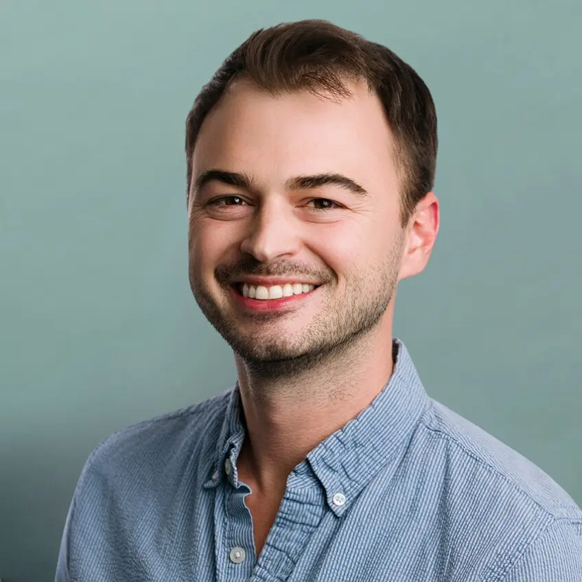 A smiling man with short brown hair and stubble wears a light blue button-up shirt, posing in front of a plain light green background.
